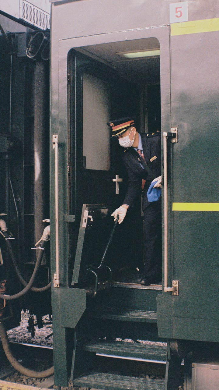 一张写实的铁路摄影。画面右侧是一节深绿色的 RW25G型软卧车厢，车身印有黄色编号 554823。两名身着蓝色制服的乘务员正站在车门旁的站台上值守。车顶的设备带有 Guangzhou (广州) 的字样。照片色调柔和，捕捉了普速列车运行中的一个静谧瞬间。A realistic railway photograph featuring a dark green RW25G (Type 25G Soft Sleeper) carriage with the yellow number 554823 on its side. Two train attendants in blue uniforms are on duty on the platform next to the carriage door. The equipment on the roof bears the name Guangzhou (广州). The photo has a soft tone, capturing a quiet moment of a conventional train in service.