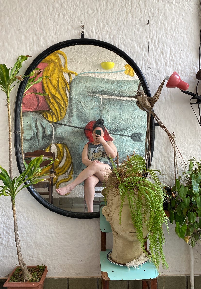 Foto de um espelho na parte externa de uma casa. A decoração ao redor é de plantas e o muro é branco. Estou em uma poltrona no reflexo.