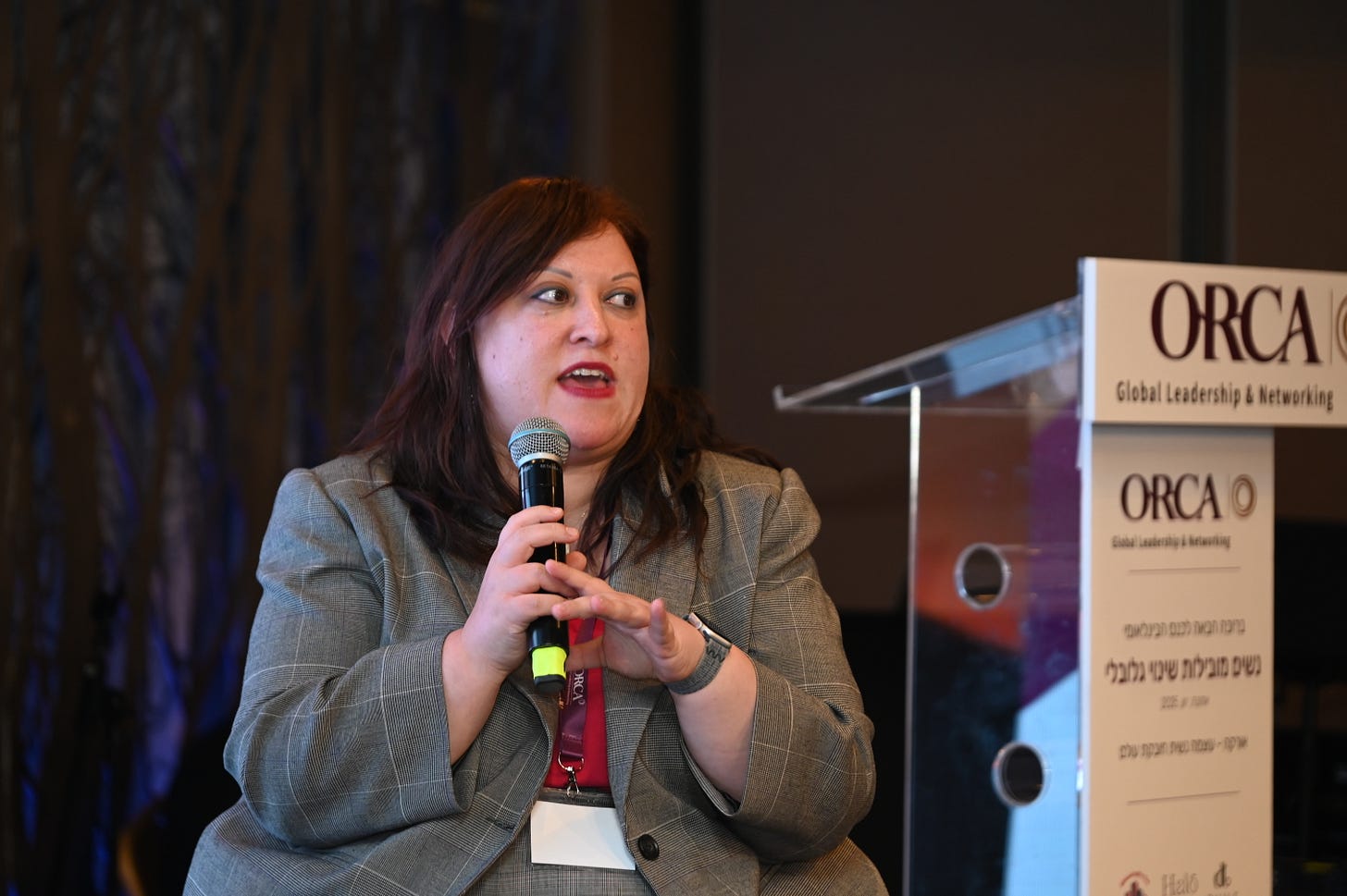 Chedva wears a grey pantsuit and pinl shirt holding a micropohone next to a podiom with the orca global logo