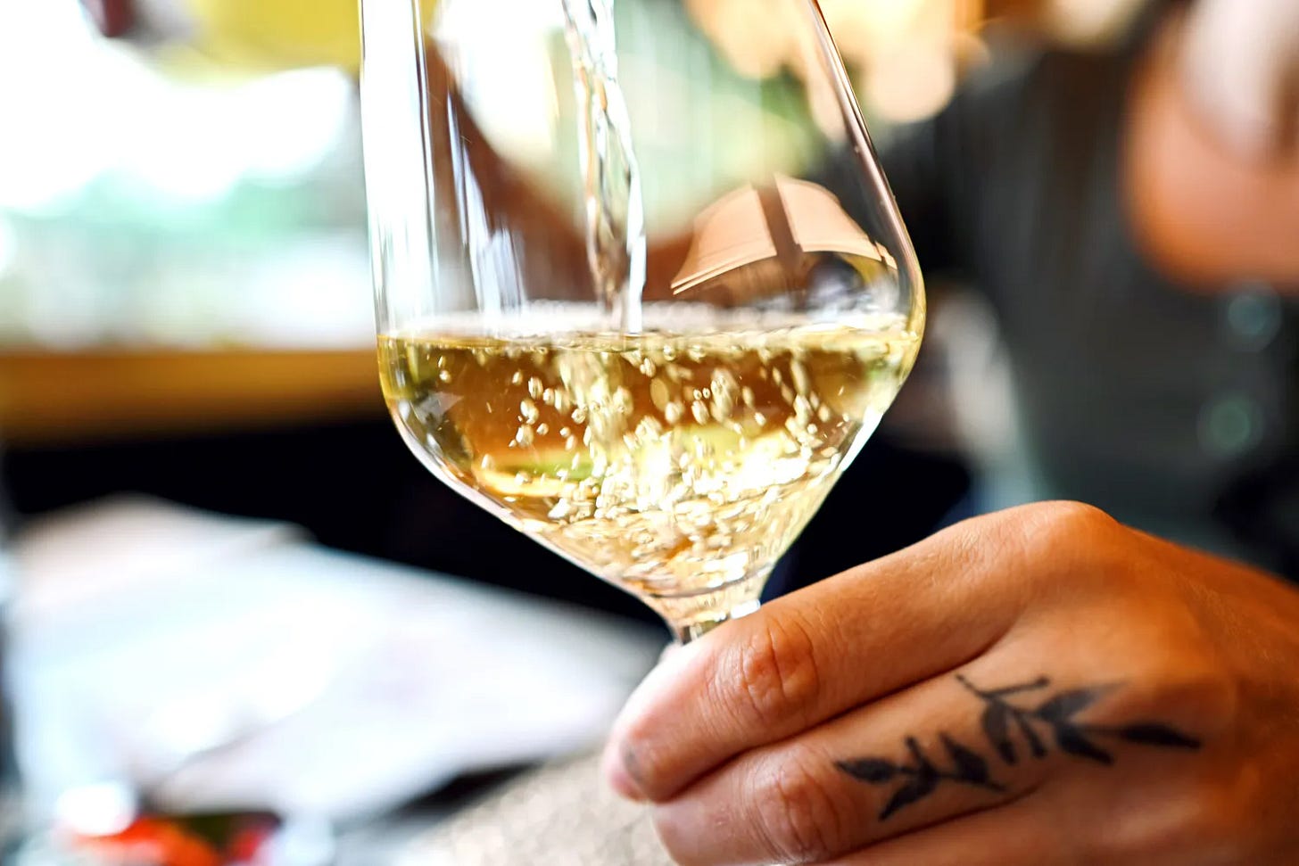 A hand with a tattoo holds a wine glass with white wine being poured