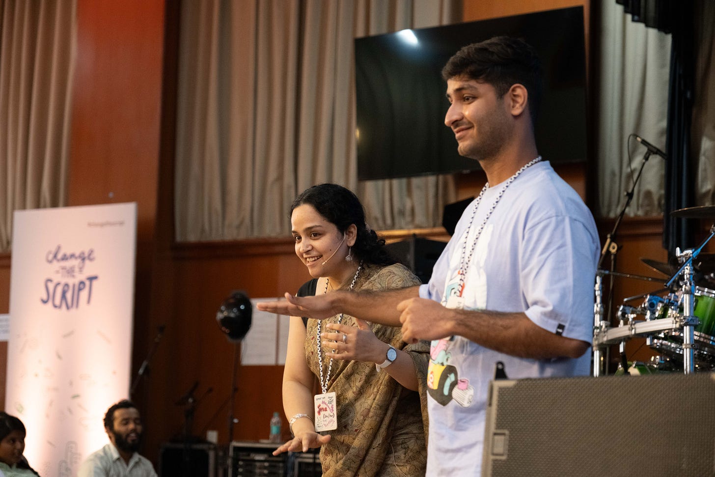 Tania is an Indian woman with long black hair pulled back. She is dressed in a beige saree with ‘kantha’ embroidery and is signing along with Parmeet Singh who is wearing a grey T-shirt. Both have taken the stage and are smiling at the audience at the Change the Script 2024 event. The stage setup includes a banner that says “Change the Script” and a visible drum kit off to the side.