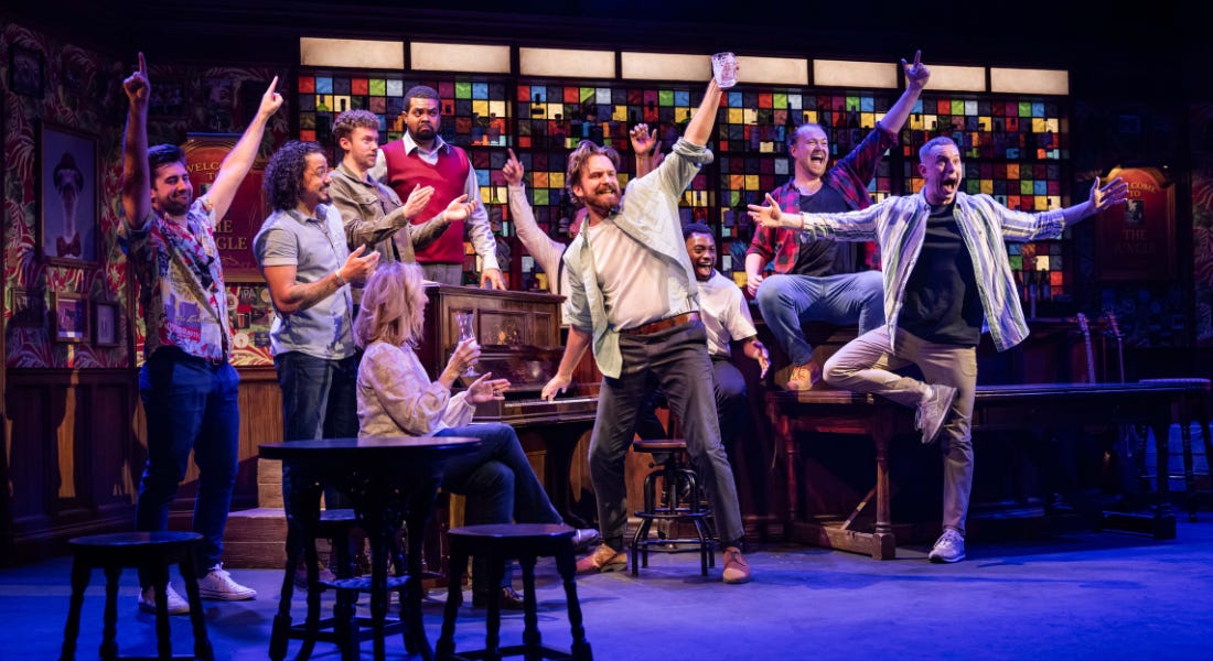 A group of men on stage holding their hands up, with a piano, stools, and colourful glass behind them A group of men on stage holding their hands up, with a piano, stools, and colourful glass behind them