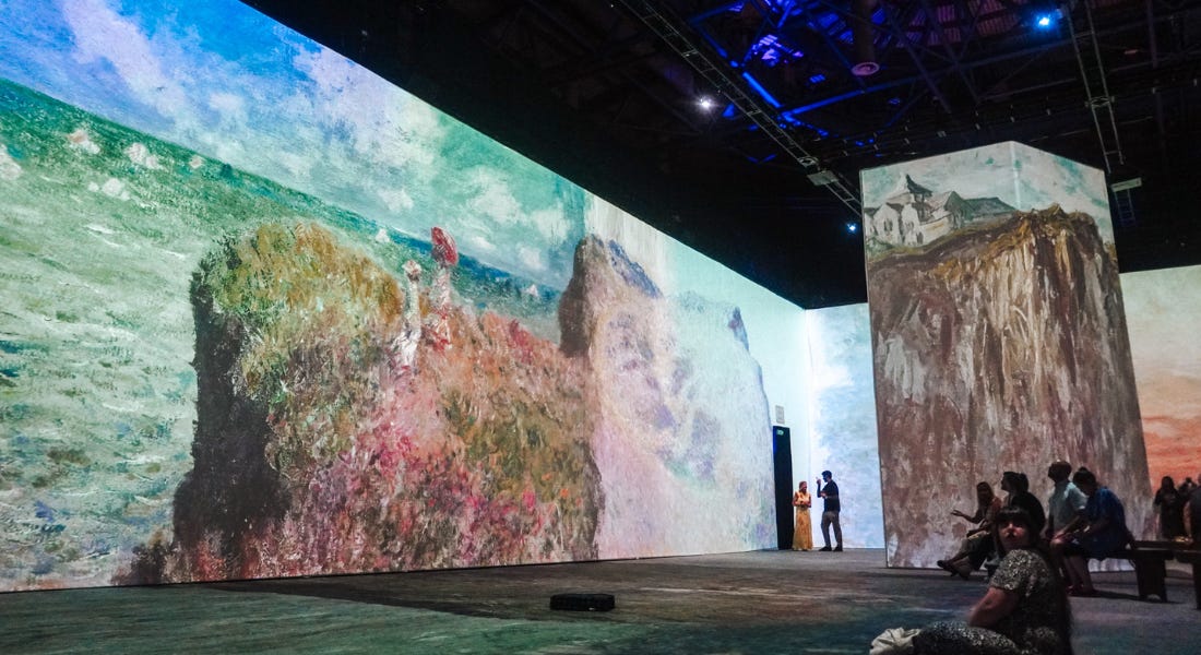 An immersive art installation with Monet paintings on the walls and floors and people sitting down An immersive art installation with Monet paintings on the walls and floors and people sitting down