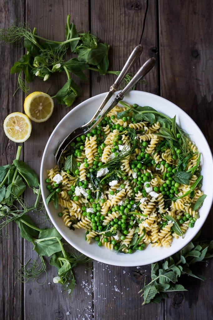 spring pea pasta salad with fresh herbs and goat cheese in white serving bowl with serving spoons. spring pea pasta salad with fresh herbs and goat cheese in white serving bowl with serving spoons.