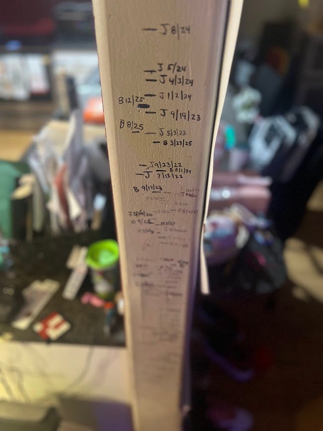 Photo of a door frame with height lines for children marked in black Sharpie with dates and letters.