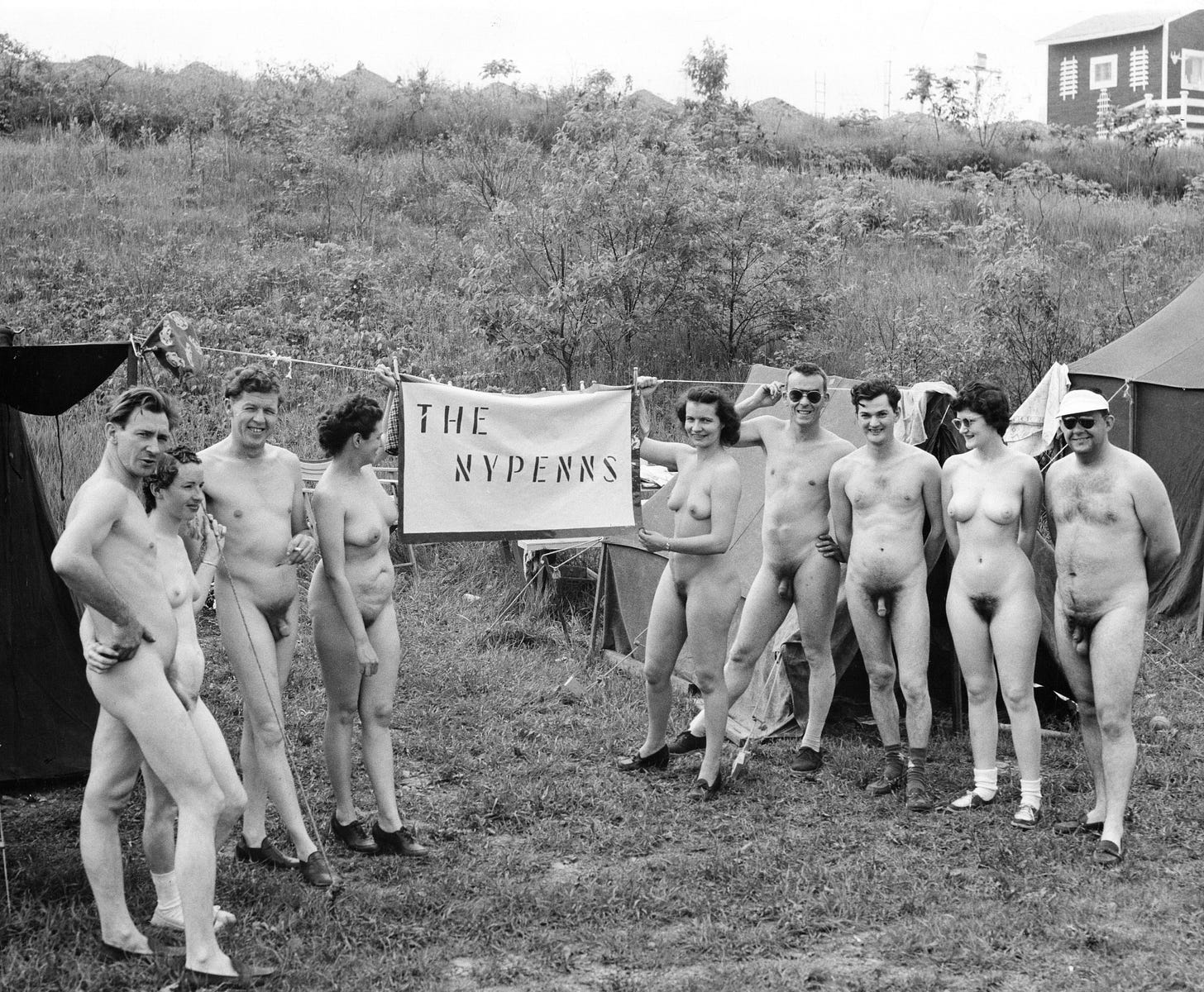 A group of nude adults standing together outdoors beside a handmade banner reading “THE NYPENNS,” with tents and a hillside in the background.