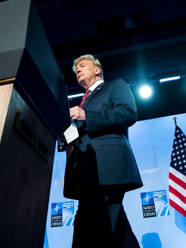 Donald Trump approaches a podium as he prepares to give a speech as president, with signs displaying the NATO logo behind him.