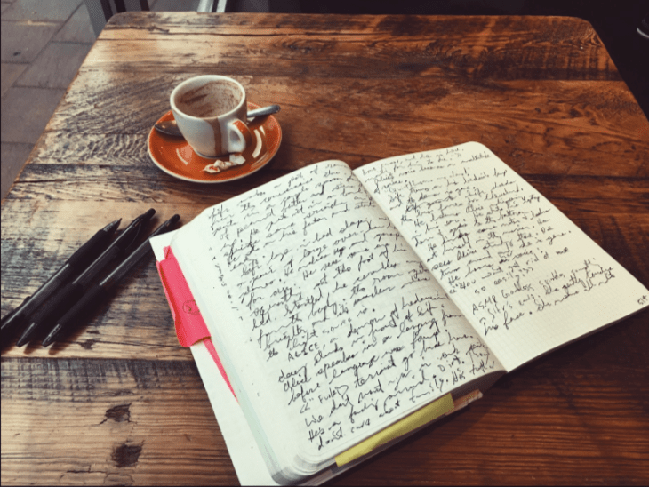 A cup of coffee, pens and an open notepad with writing in it on a wooden table