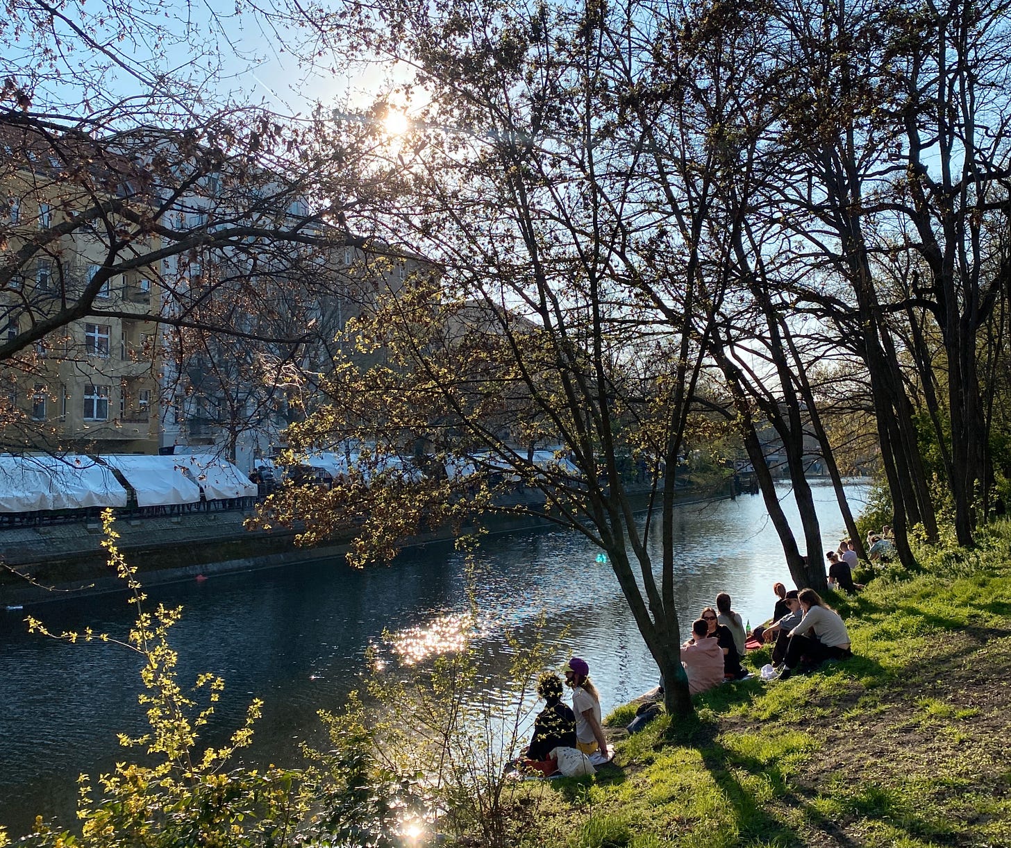 Foto de um canal de água com pessoas sentadas na grama próxima à água. Árvores acima delas e prédios residenciais na frente, sob um céu azul e ensolarado, em Berlim.