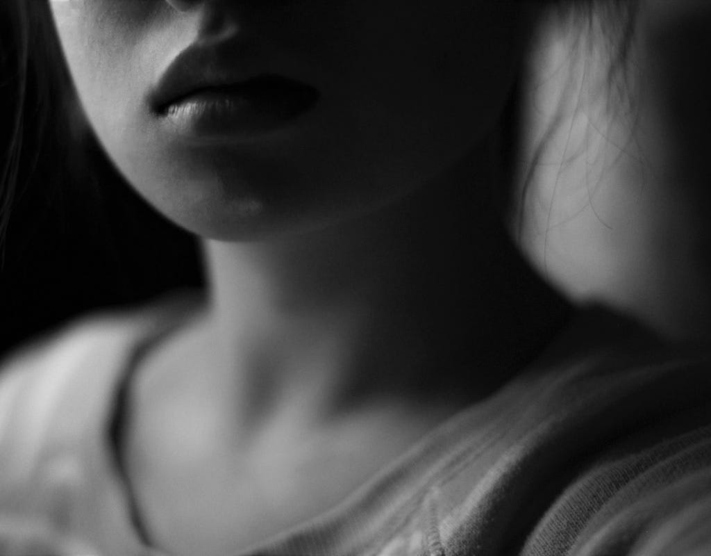 A young woman with dark hair pulled back with a few long wisps of hair framing either side of her unsmiling face. Only her full lipped closed mouth, chin and upper torso clothed in a tank top shows in this black and white photo. 
