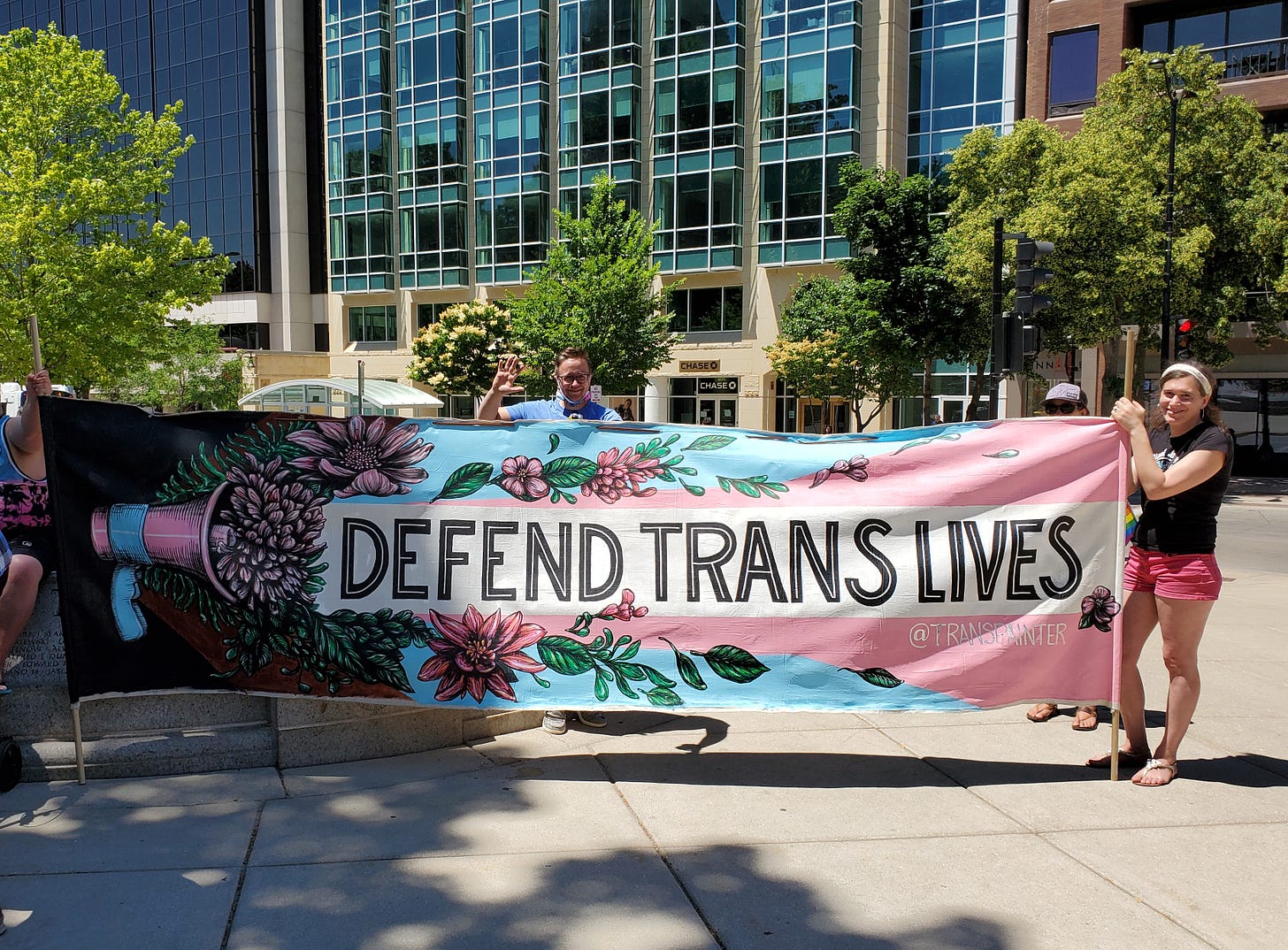 Two people stand and hold a large banner that reads "Defend Trans Lives" and is illustrated with bold and colorful flowers sprouting from a megaphone. They are outdoors in warmer weather.