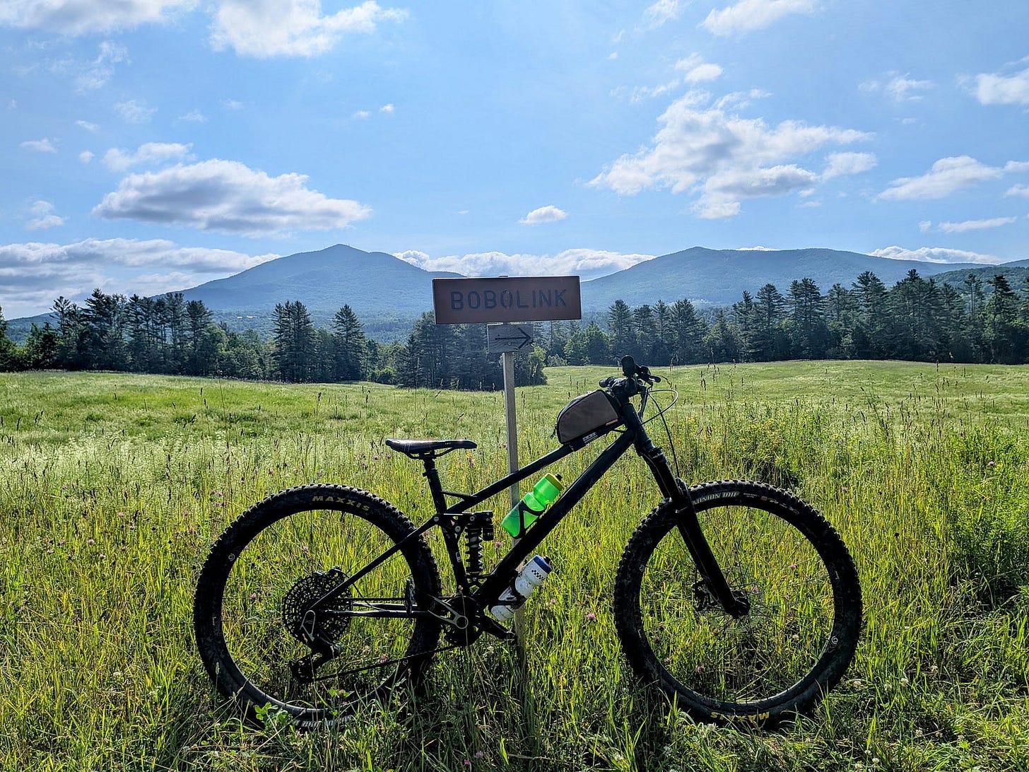 A black full-suspension mountain bike with green water bottles parked in a grassy meadow next to a brown trail sign reading Bobolink, with forested hills and distant blue mountains under a partly cloudy sky A black full-suspension mountain bike with green water bottles parked in a grassy meadow next to a brown trail sign reading Bobolink, with forested hills and distant blue mountains under a partly cloudy sky