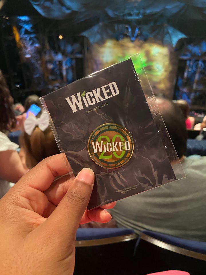 From top left to bottom right - going across horizontally. Image 1) Alexa is holding up a green frisbee that says Defying Gravity since 2003 in black font. The text is curved around the frisbee, in a half circle. In the center, there is a black stenciled picture of Elphaba holding up her Broome. In the middle of skirt, the text “Wicked” is printed with a 20 in the background of the word. Both ‘Wicked’ and the number ’20’ are highlighted in green font. Image 2) The back of Alexa’s left hand. She has a temporary tattoo printed that reads “Toss Toss” in red cursive ink. The two T’s are connected, and there is an asterix/flower type symbol over the second to last S. In smaller font, “Wicked” is printed in all caps. Image 3) Alexa is posing in front of a replica of the clocktower, as seen in the Wicked production. At the top of the clock tower, which is wooden, with antique looking wheels and roman numerals, there is a dragon neck (with its head cut off in the photo) with webbed looking wings. Alexa posing side wise, with a surprised expression on her face, as if she is being scared by someone. She is wearing a polka dotted scarf in her hair, and a pink collared shirt (you can only see her from the shoulders up.) Image 4) Alexa is posing with Carrie St. Louis, who played Glinda on Broadway! Carrie is on the left, wearing a pink satin looking pant suit with a tie around the blazer. Her blond hair falls around her collar bone. She is smiling. Alexa is wearing the same outfit as she was in the previous photo. You can also see her jeans in this photo, along with a pink pin on her shirt. Carrie and Alexa are standing in front of pink background, blocking text that says “it’s good to see me, isn’t it?” Image 5) Alexa is holding up a pink gift bag that reads “Let’s have a celebration the Glinda Way” and the Wicked 20 logo. You can see two pink satin scrounges and a bracelet with pink glass beads on her wrist. You can also make out a few audience members in back of the bag. Image 6) Alexa is holding a light pink baseball cap with the Wicked 20 logo on the front. The text, Wicked, is in White, while the 20 is in green. The entire logo is outlined in black. You can see audience members again, and a little bit of the stage. Image 7) Alexa is holding a pin with the Wicked 20 logo. Wicked is in white, and 20 is in green. Both are outlined in gold. The base of the pink is a golden color, and the outline of the pin has roman numerals like a clock face - in dark green. Image 8) The back of two women, wearing two of Glinda’s iconic costumes - the pink, strapless, frilly, sparkly, short dress from Dancing through Life (left) and the blue dress from the top of the show (right) The blue dress is longer for the show, but this woman’s dress is a bit shorter.