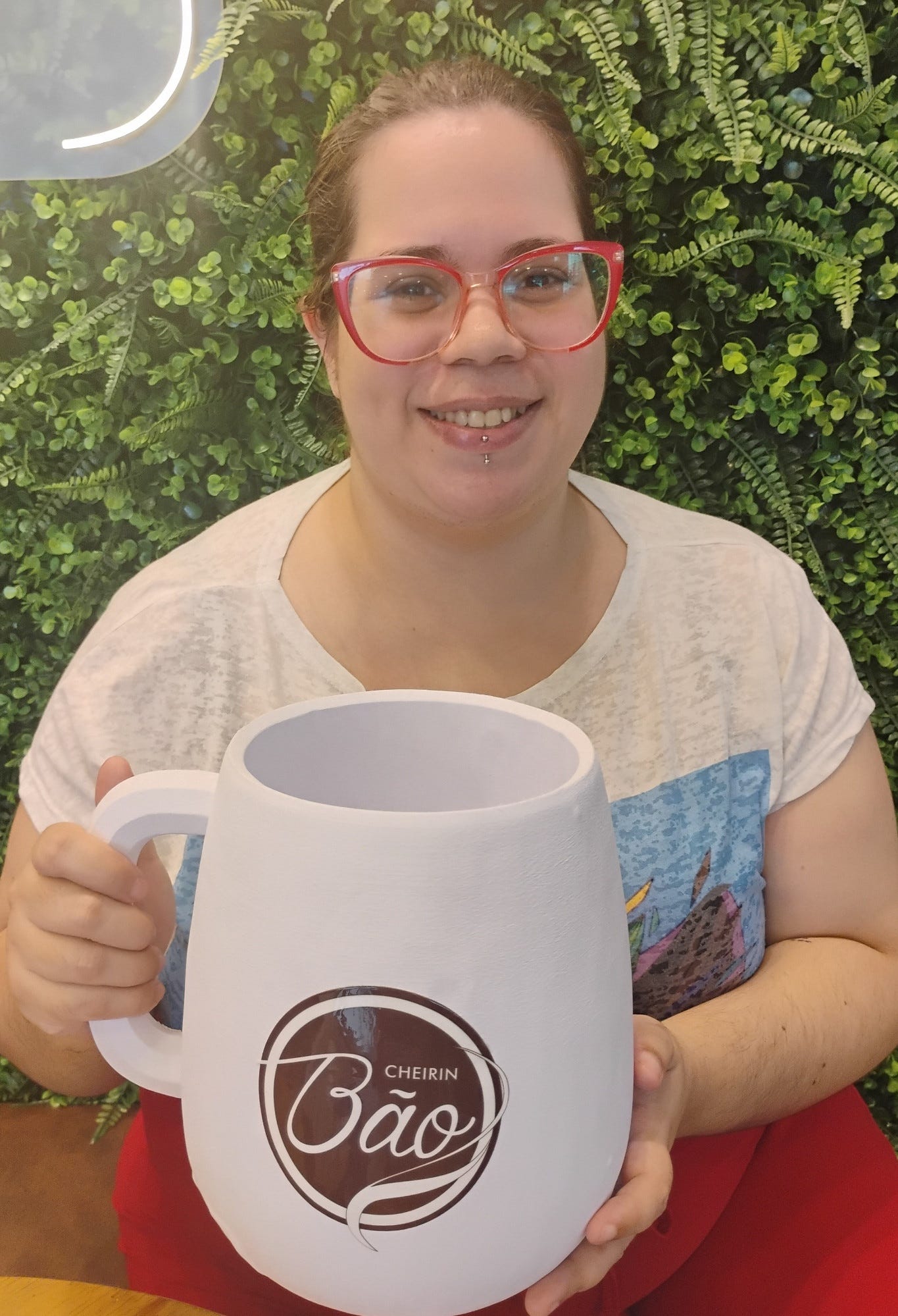 A imagem mostra uma mulher sorridente segurando uma caneca branca gigante personalizada com o logotipo da rede de cafeterias Cheirin Bão. A mulher usa óculos com armação vermelha e cabelos presos, vestindo uma camiseta branca e calça vermelha. Ela segura com as duas mãos uma caneca de tamanho exagerado, que funciona como um item promocional ou decorativo da marca. O logo "Cheirin Bão" está centralizado em um círculo marrom. O Ambiente: O fundo é composto por uma parede de folhagens verdes (jardim vertical).