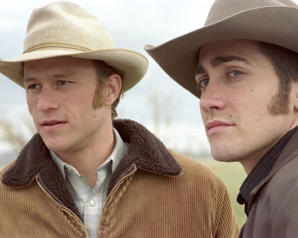 Two men in cowboy hats and brown jackets look off into the distance. 