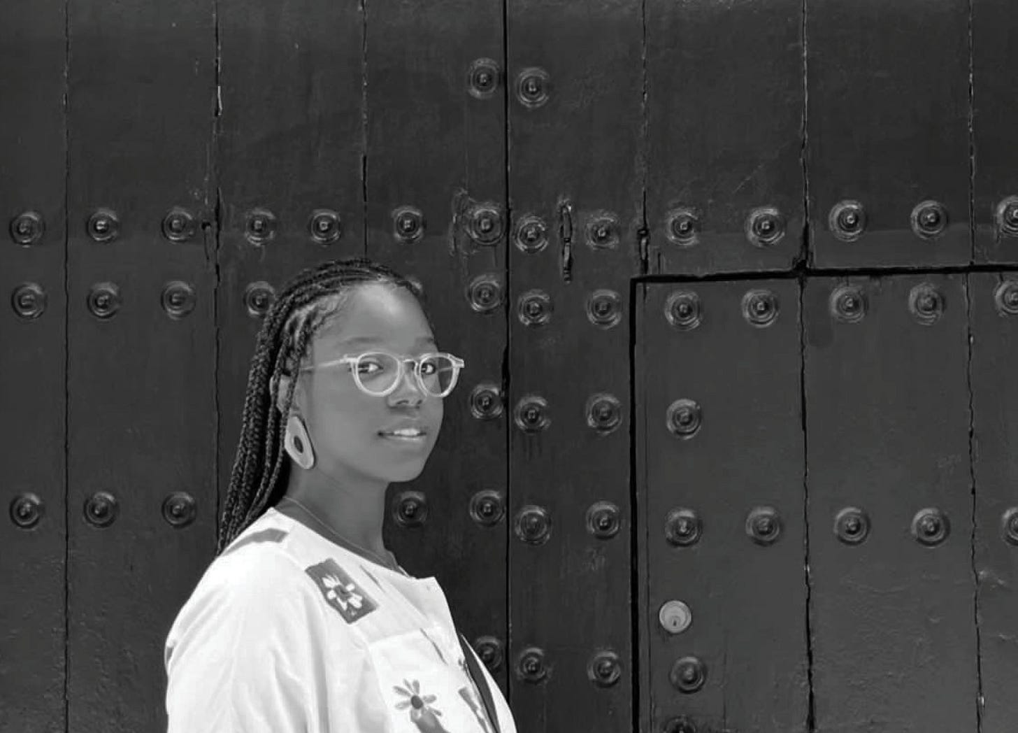 Image Description: Black and white photo of Ravon Ruffin Feliz standing in front of a black background and door wearing Fulani braids, a white shirt featuring flowers and a pair of clear eyeglass frames while staring directly into the camera with a soft gaze and even softer smile. This photo was taken by their wife on their honeymoon last year while in Spain. Image Description: Black and white photo of Ravon Ruffin Feliz standing in front of a black background and door wearing Fulani braids, a white shirt featuring flowers and a pair of clear eyeglass frames while staring directly into the camera with a soft gaze and even softer smile. This photo was taken by their wife on their honeymoon last year while in Spain.