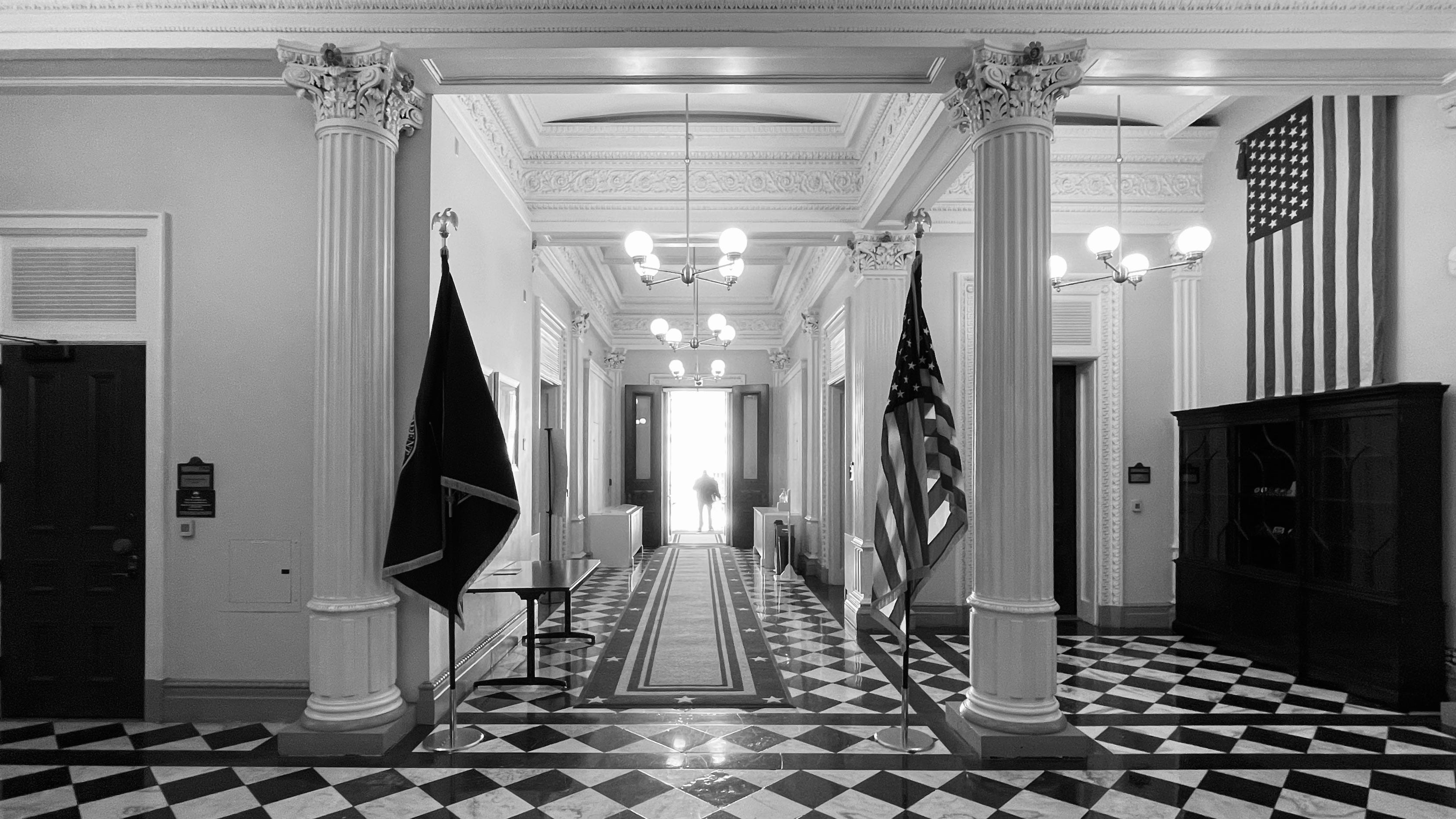 Black and white photo of a person standing outside an open door at the end of a short hallway at the Eisenhower Executive Office Building, looking out toward the White House.