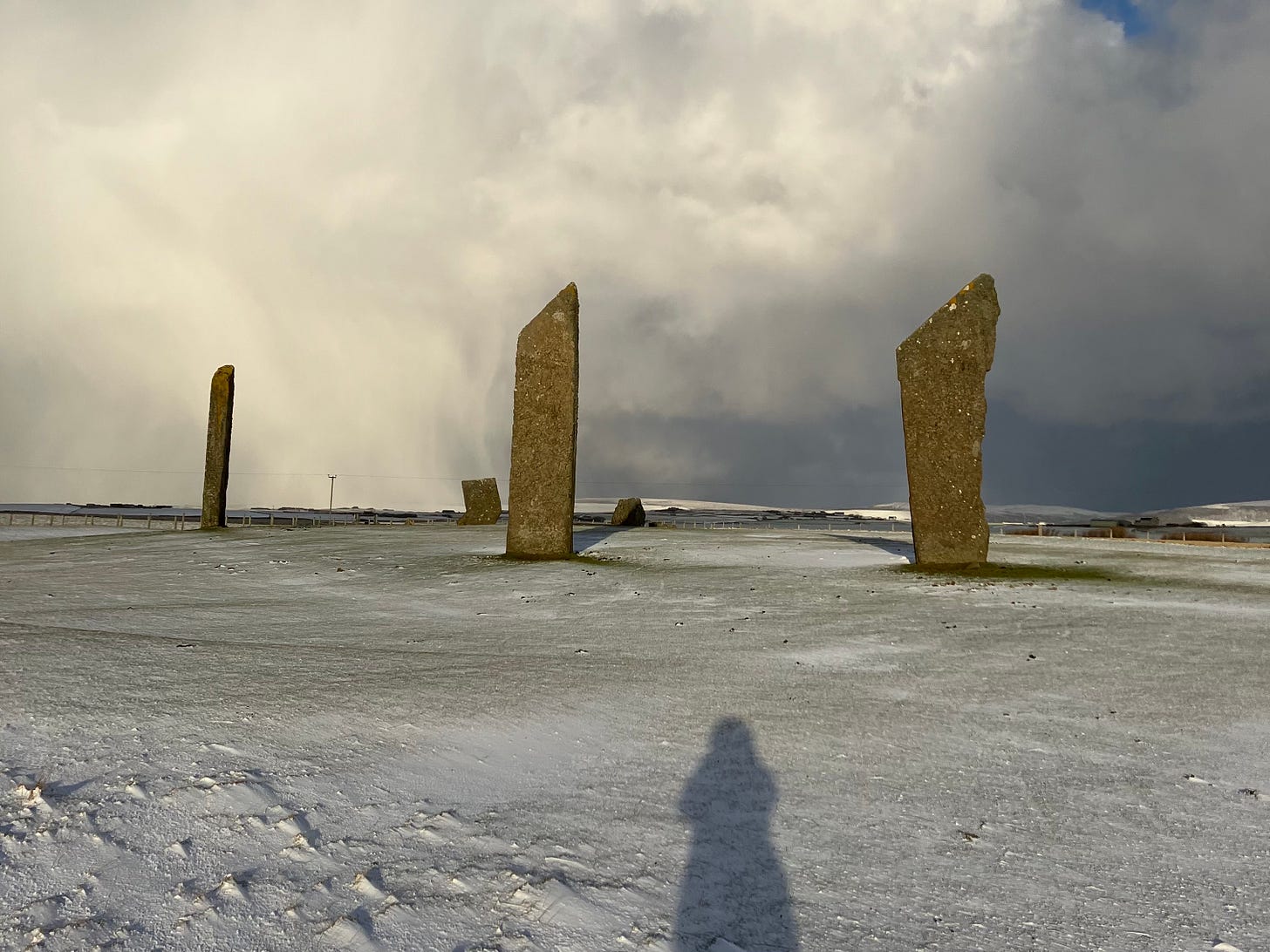 A winter landscape with white snow covered expanse broken by three very tall menhirs and the shadow of the photographer (me) stretching toward them.  The horizon is filled with billowing white clouds. 