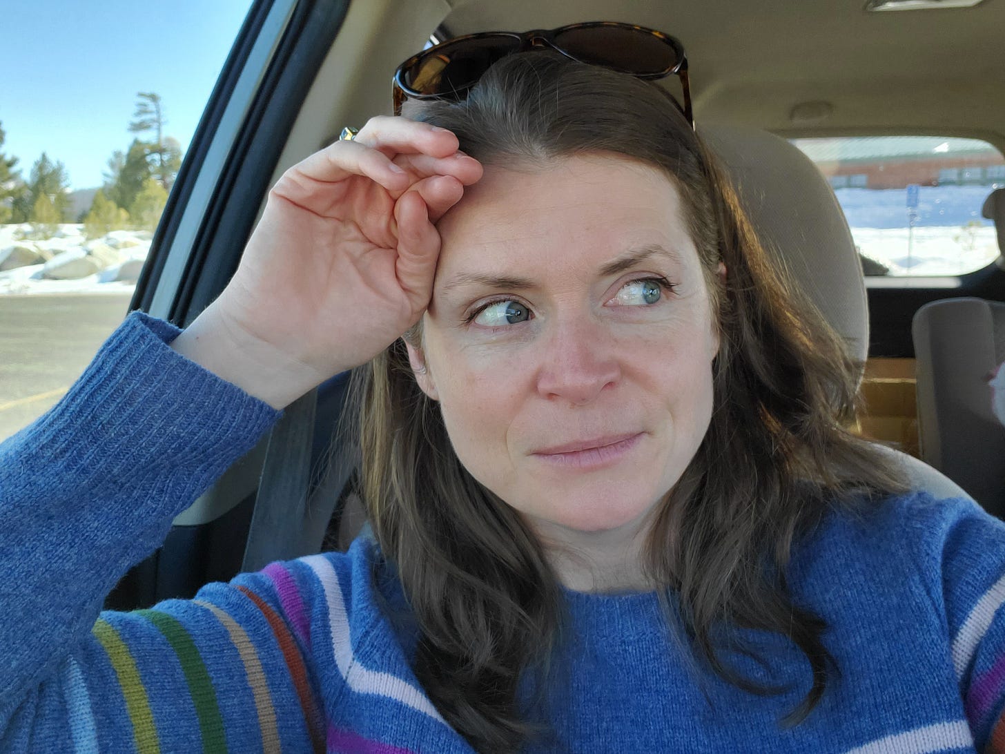 Image of a woman in the driver's seat of a car with her elbow on the open windowsill, sunglasses on her head, and quietly looking toward the passenger side of the car.