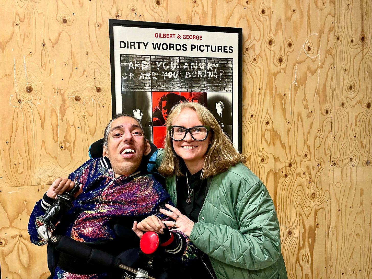 Image Description 4: In front of a film poster on a wooden wall at Central Saint Martins, closeup portrait photo of Sulaiman (a wholeheartedly Disabled AF and British-Pakistani man with black buzzcut hair and features “white hair of wisdom”) sitting  in his power wheelchair next his friend Deborah (a badass white woman with shoulder-length blonde hair and large black framed glasses. The top of the film poster can be seen just over their heads of an image of a stone wall with all caps words  saying, “Gilbert & George - Dirty Word Pictures - Are you angry or are you boring?” Sulaiman is wearing his joyous sequinned casual jacket. Deborah is wearing a knee-length pale green down coat and a long black dress. Both, Sulaiman and Deborah, are smiling and joyful. Descriptions END. 