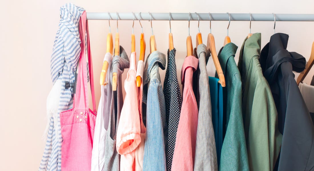 A clothing rail with colourful shirts and bags hanging from it, with a light pink wall behind it A clothing rail with colourful shirts and bags hanging from it, with a light pink wall behind it
