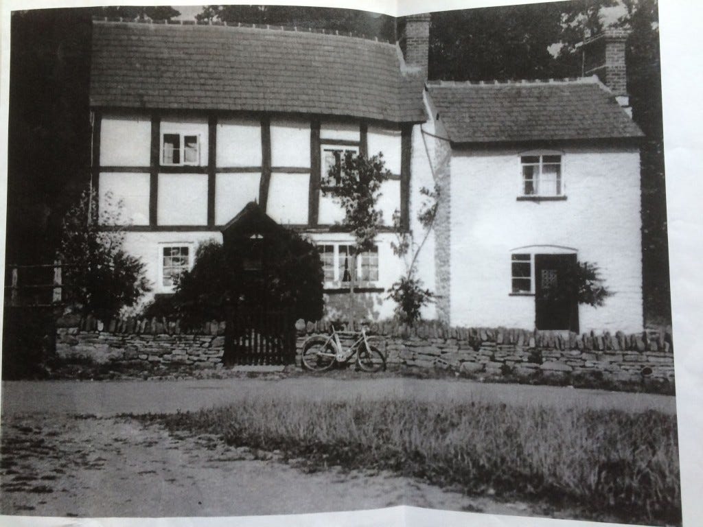 Black and white photo of the cottage looking much neater than it does right now. There’s an old-fashioned bicycle resting in front of the stone wall by the gate.