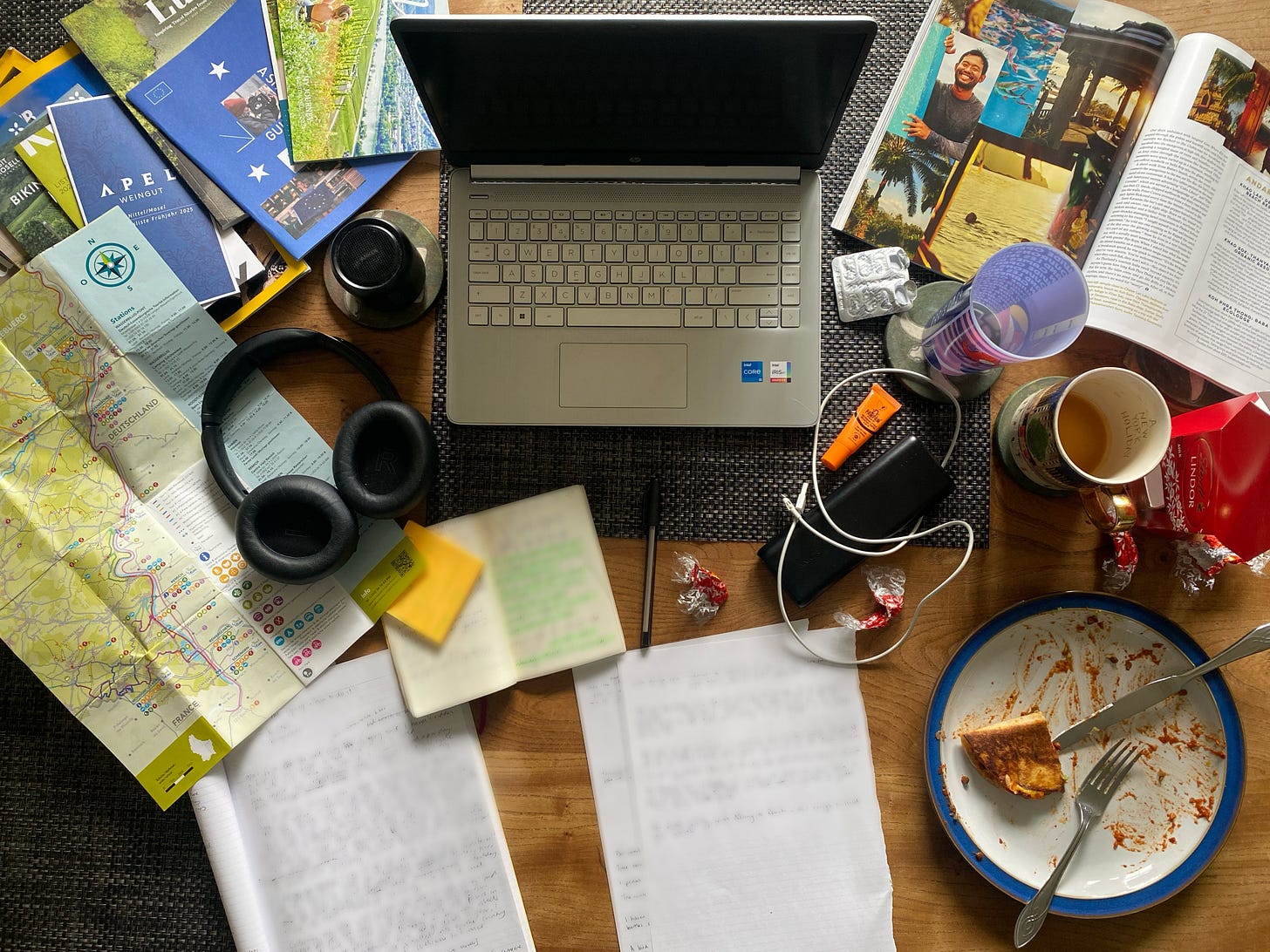 A cluttered travel workspace with an open laptop, maps, notebooks and headphones, alongside a mug of tea and a half-eaten taco A cluttered travel workspace with an open laptop, maps, notebooks and headphones, alongside a mug of tea and a half-eaten taco