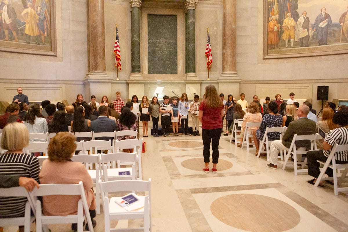 in the foreground, people are seated in folding chairs in the Capitol rotunda. In the distance, a line of children stands in front of two American flags. in the foreground, people are seated in folding chairs in the Capitol rotunda. In the distance, a line of children stands in front of two American flags.