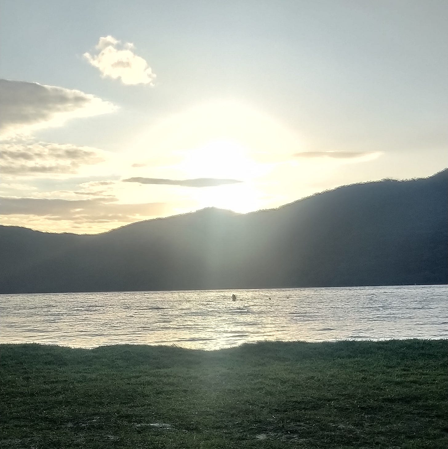 Foto colorida em local aberto da Lagoa do Peri, em Florianópolis. A lagoa está com a água bem calma, e o sol reflete nela deixando tudo dourado e aconchegante. O sol está se pondo atrás de um morro lá no fundo, sua cor apenas a sombra no reflexo do sol. Uma pessoa, minúscula no fundo da lagoa, se banha e aproveita o momento.