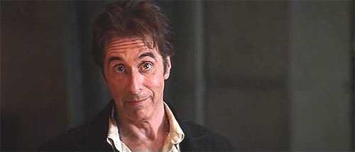 Gif dal film "L'avvocato del diavolo" con Al Pacino che sorride e tira su le mani con gli indici puntati verso l'alto. Sfondo grigio non a fuoco.