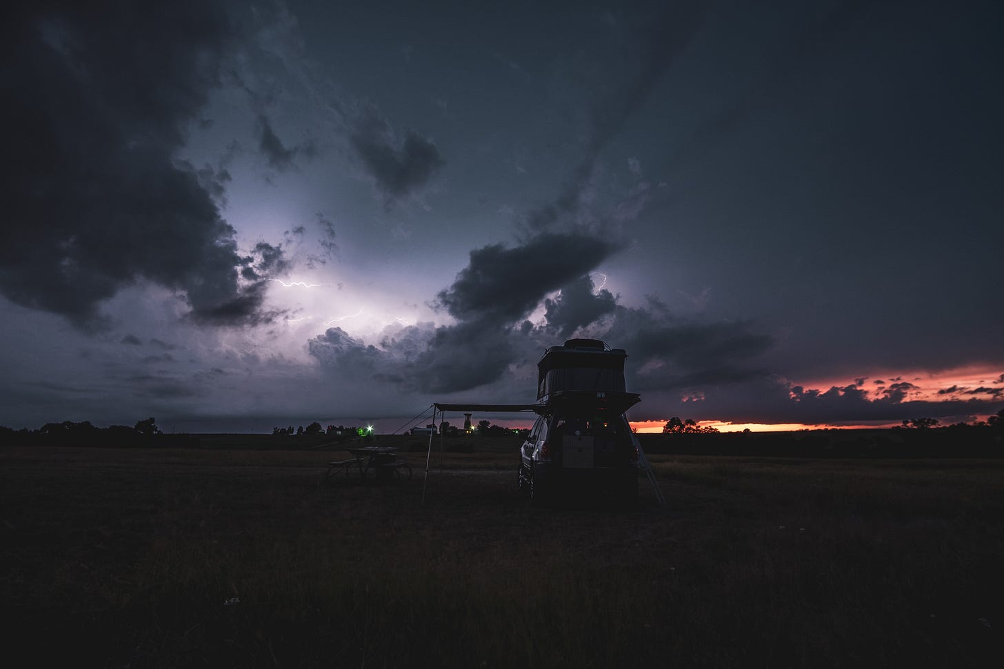 The dark-colored Ford Escape is parked in a grassy field in Nebraska at sunset, with its rooftop tent open and set up. The scene is lit by the warm, golden light of the setting sun, and lightning is visible in the dramatic sky.