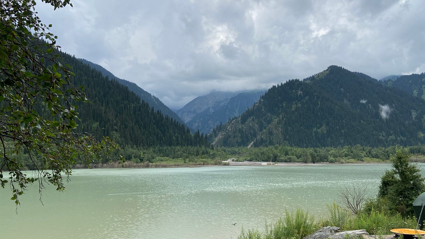 Lake Issyk in Kazakhstan. It is a seafoam-colored lake surrounded by tree-covered mountains. The sky is overcast.