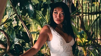 A woman in a white dress stands in the shade of plants on a tropical island