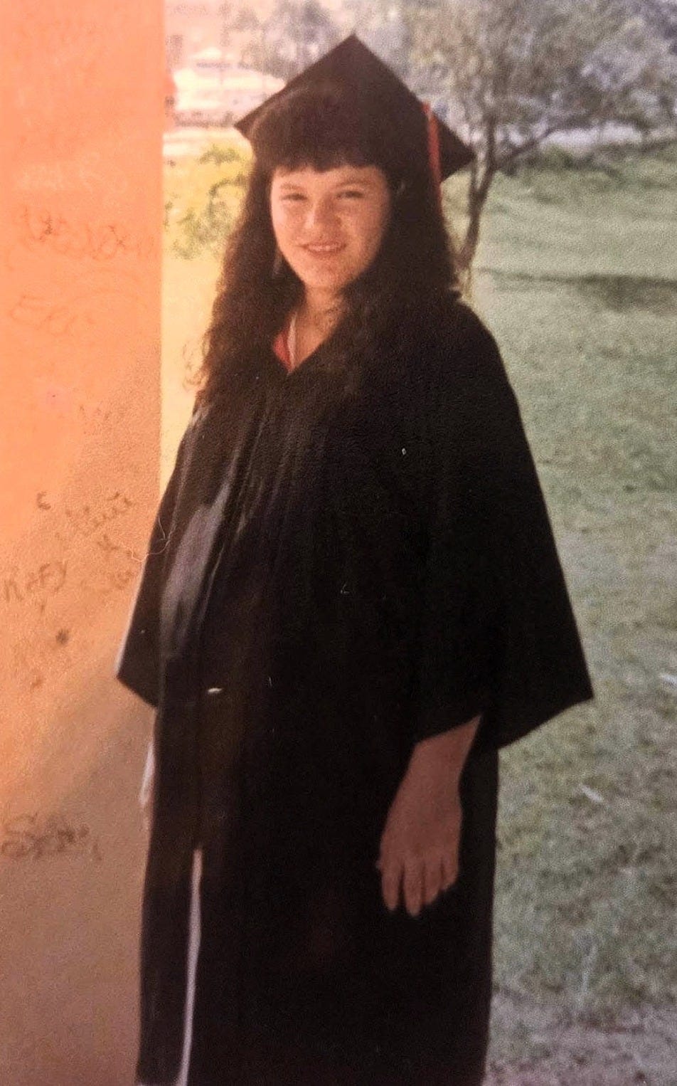 A woman wearing a black graduation cap and gown stands outdoors. She is visibly pregnant. The photo quality is old and faded.