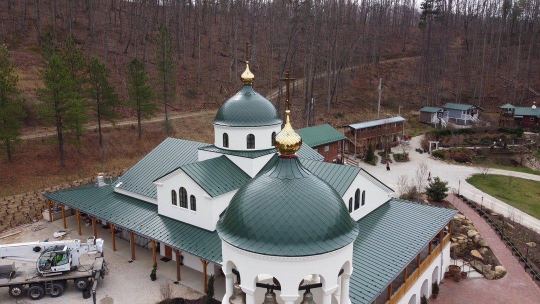Aerial view of a church under construction with green metal roofing, gold-topped domes and crosses, a crane on site, surrounded by bare winter trees.