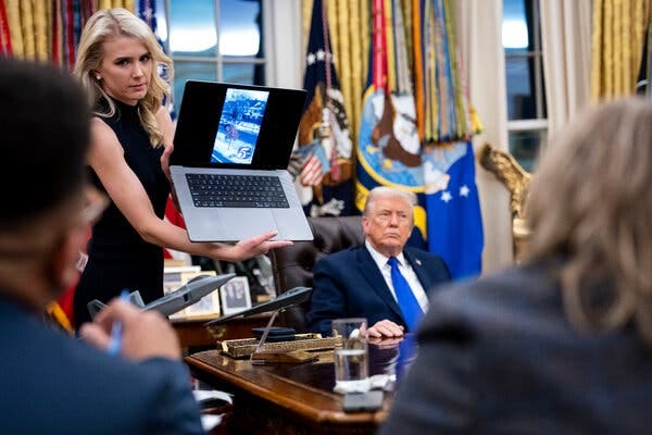 A woman holding a laptop while President Trump is seated at the Resolute Desk in the Oval Office.