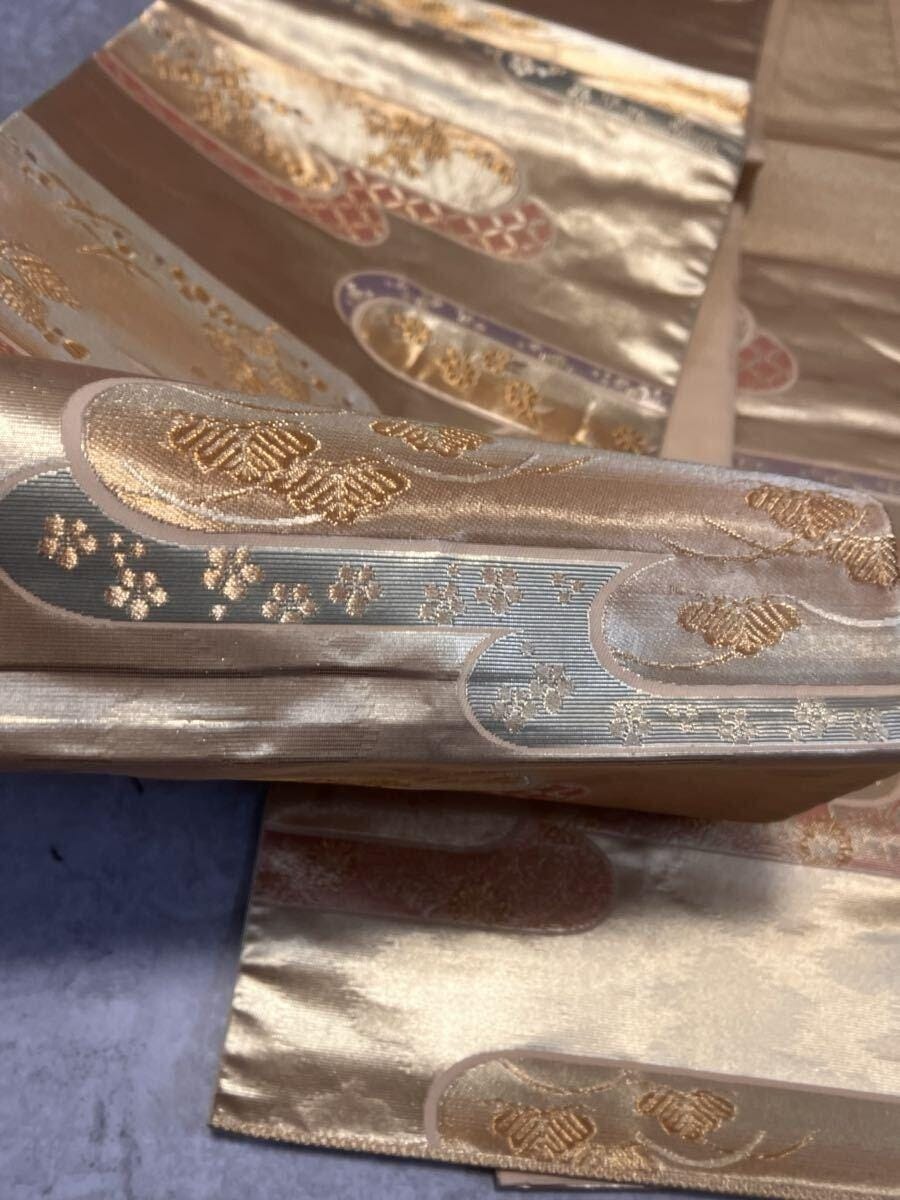 A formal fukuro obi in gold kinran brocade, folded for presentation — the streaming cloud motifs and scattered paulownia crests worked in raised gold thread against a lustrous gold ground. The weight of the metallic thread holds the silk in its own architecture. A formal fukuro obi in gold kinran brocade, folded for presentation — the streaming cloud motifs and scattered paulownia crests worked in raised gold thread against a lustrous gold ground. The weight of the metallic thread holds the silk in its own architecture.