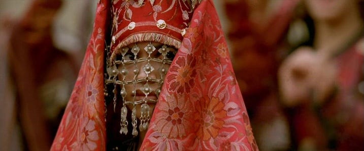 Rosario Dawson wedding veil in Alexander – Intricate red and gold wedding veil with beaded face covering from Alexander Revisited: The Final Cut. Note: Still shown is promotional and not taken directly from the 4K UHD.