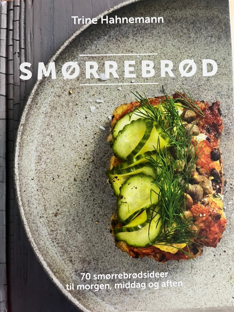 The cover of the book Smørrebrød by Trine Hahnemann. The cover photo shows an open-faced sandwich with cucumber slices, a red sauce, and a green herb, on a round, gray plate, on a gray wooden table