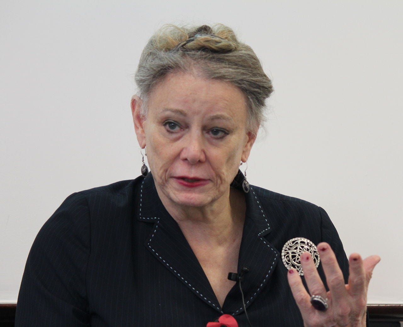 Deirdre McCloskey wearing a dark striped suit top, gesticulating with left hand, appearing to speak, while looking right of camera Deirdre McCloskey wearing a dark striped suit top, gesticulating with left hand, appearing to speak, while looking right of camera
