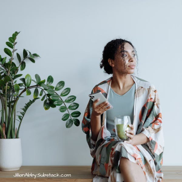 A woman with curly hair sits on a wooden bench beside a leafy green plant, holding a smartphone in one hand and a glass of green juice in the other. She is wearing a light blue top and a floral-patterned robe, gazing thoughtfully out of frame. The background is a minimalist white wall with the text "JillianAbby.Substack.com" in the lower left corner.