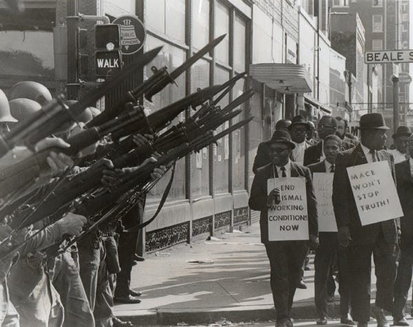 national guard soldiers poise their guns in front of Black protestors holding signs reading "mace won't stop truth!" and "end abysmal working conditions now"