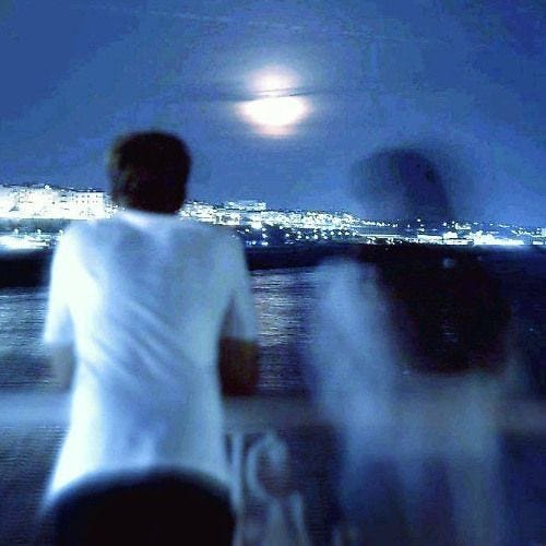 This may contain: two people standing on a boat looking out at the water and city lights in the distance This may contain: two people standing on a boat looking out at the water and city lights in the distance