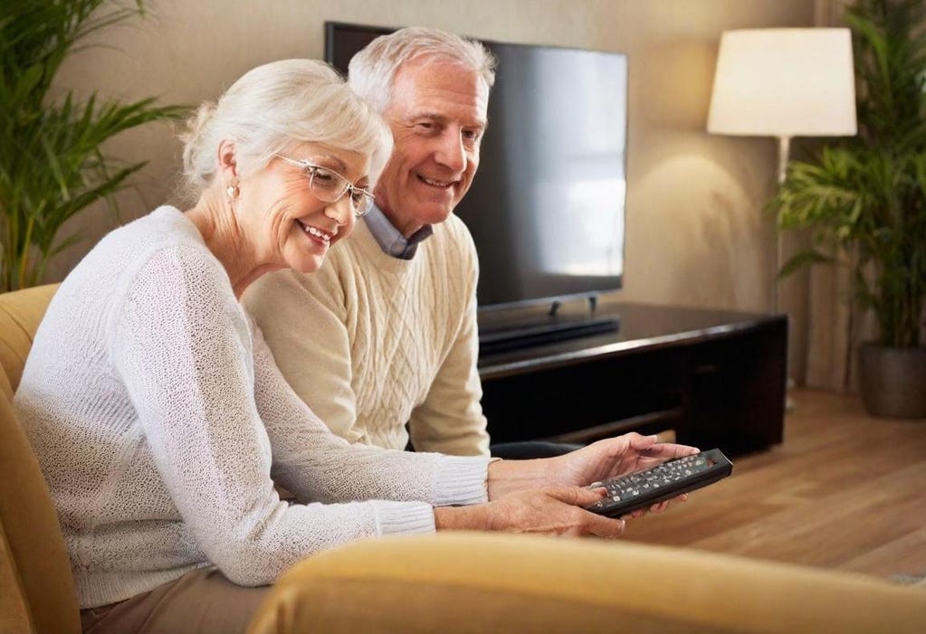 Portrait of a 75-year-old white couple with white hair sitting on a couch with their TV in the background. Nearest to the camera is the wife, who is holding a TV remote. Both are smiling and looking off-camera to the right of the photo.  