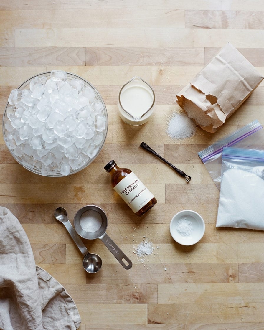 a glass bowl of ice, a small bottle of vanilla extract, measuring spoons an other cooking items laid out on a wooden table