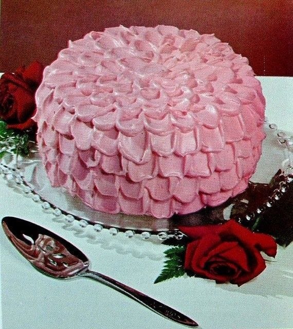 This may contain: a pink frosted cake sitting on top of a table next to two red roses