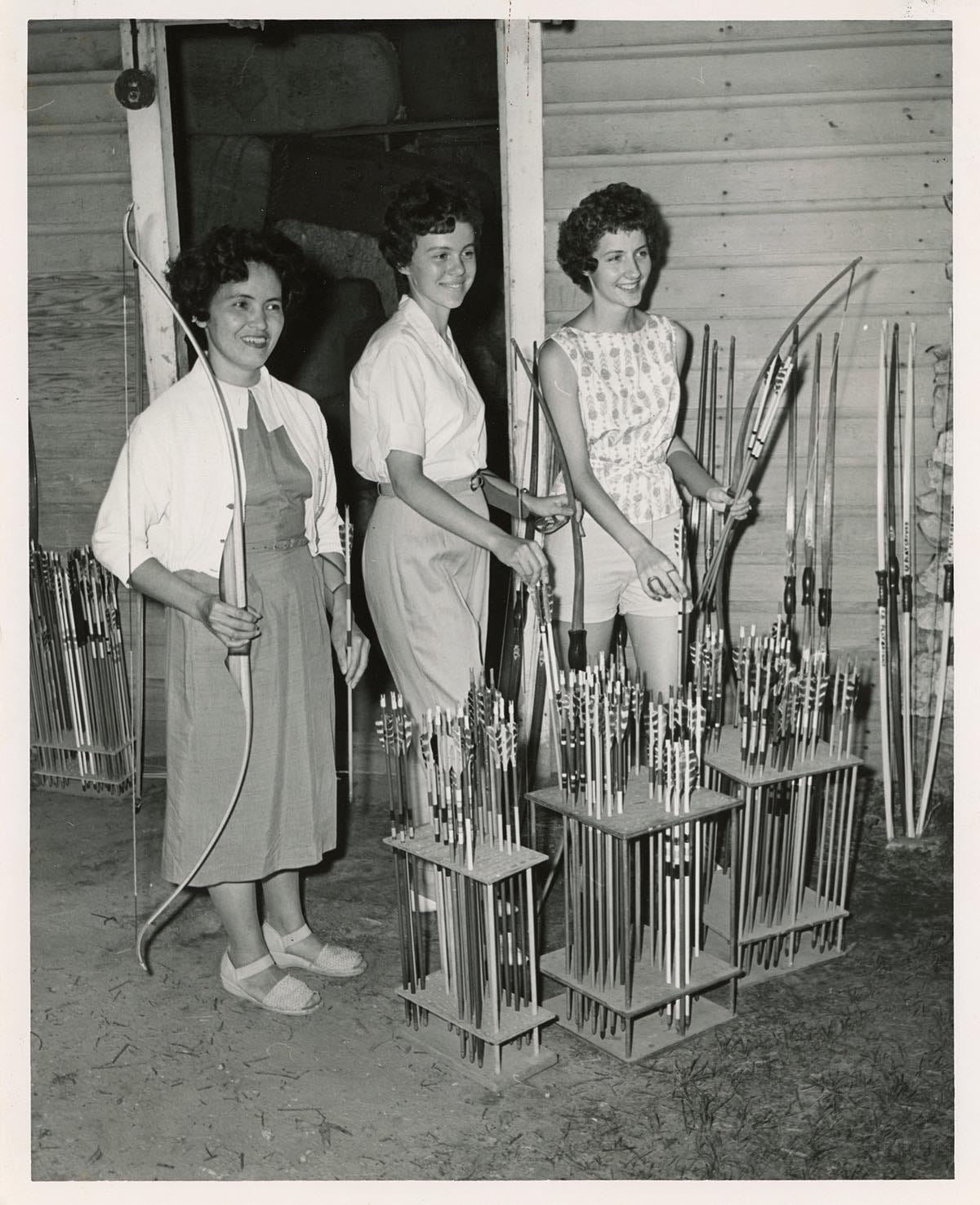 three white women in 1960s fashions pose with archery equipment, with stands of arrows in front of them. Karrie's mother is on the far right. 