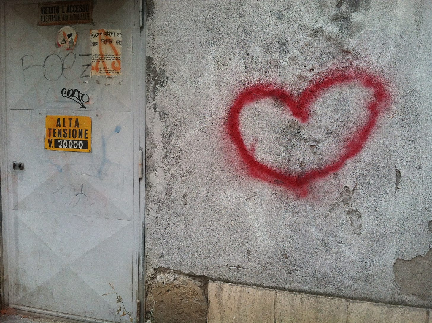 a color photo of a graffiti heart on a wall next to a door upon which is a sign that says "ALTA TENSIONE V. 20,000"