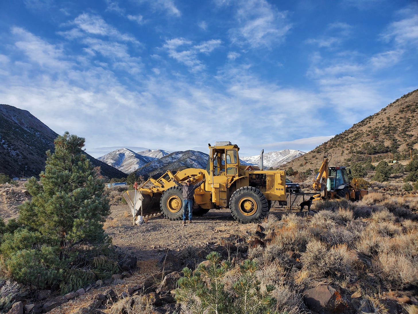 Eric spreading his arms out wide in front of a large front loader tractor. White Mountains in the distance.