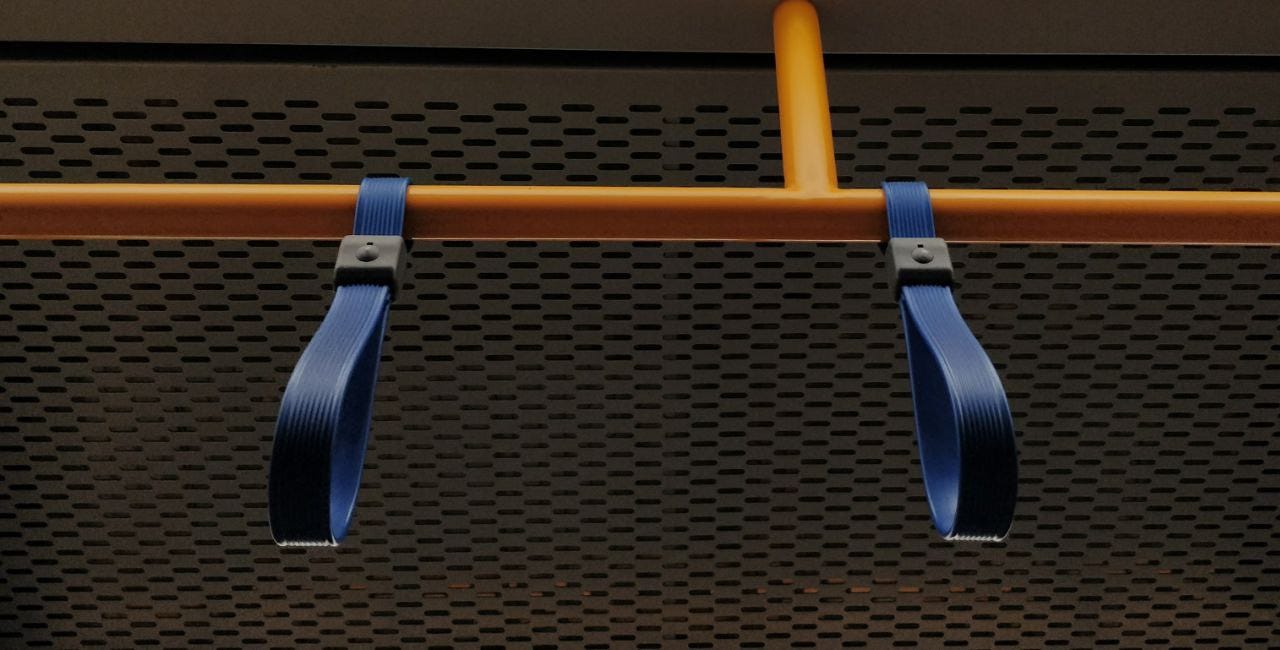 Sfondo grigio bucherellato del soffitto di una metropolitana visto dal basso con un tubo giallo che corre orizzontalmente e due maniglie in plastica blu.